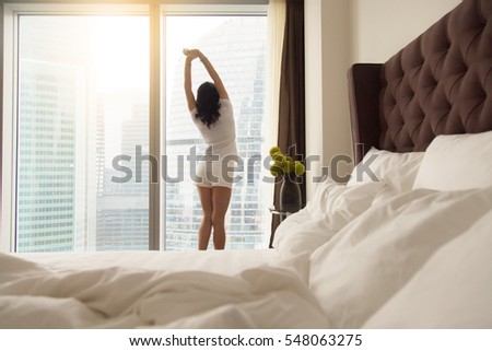 Unmade bed with white pillows and sheets in cozy bedroom, stretching young model looking in window at city view, temptation to sleep more, ready to rise, healthy day start, exercising to get energy