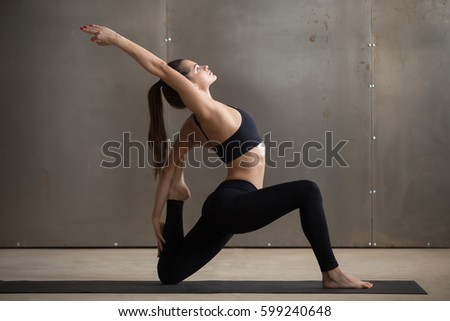 Young attractive woman practicing yoga, standing in Horse rider exercise, anjaneyasana pose, working out, wearing black sportswear, cool urban style, full length, grey studio background, side view