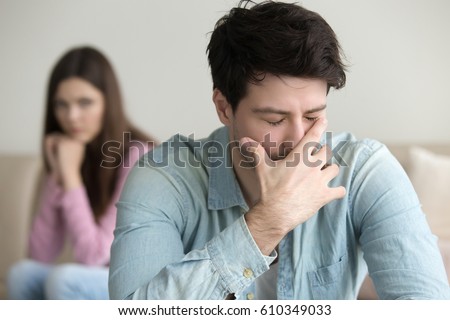 Young guy is very upset and crying, feeling guilty, depressed man in pain, girlfriend sitting in the background indoors, family relationships problems concept, businessman having troubles at work
