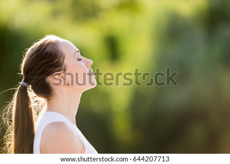 Calm beautiful smiling young woman with ponytail enjoying fresh air outdoor, relaxing with eyes closed, feeling alive, breathing, dreaming. Copy space, green park nature background. Side view portrait