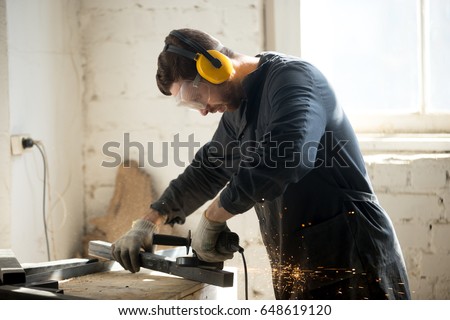 Worker in workwear, protective glasses, hearing protection headphones, gloves using dangerous electric power tools in loft workshop. Safety at workers workplace, personal protective equipment concept