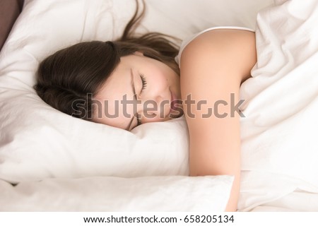 Young woman napping while hugging soft pillow, resting in cozy bed with fresh white sheets, smiling in her sleep. Healthy sleep, deep relaxing and strength renewal after hard work week concept