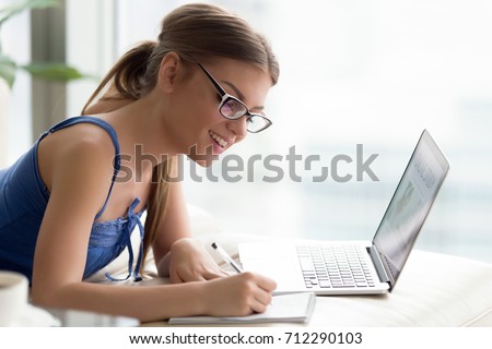 Happy young woman wearing glasses studying online on laptop, writing down in notebook important information from course, student choosing e-learning program, easy effective self education, side view
