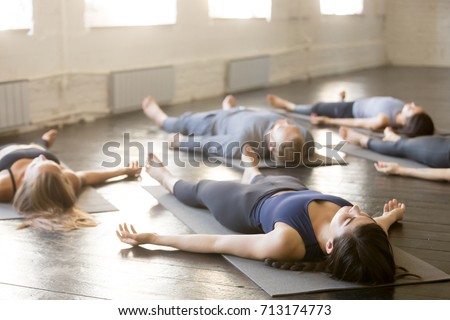 Group of young sporty people practicing yoga lesson in gym, lying in Corpse exercise doing Savasana pose, friends relaxing after working out in sport club, indoor image. Wellbeing and wellness concept