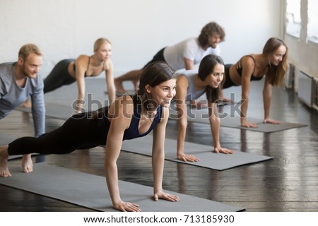 Group of young sporty attractive people practicing yoga lesson with instructor, doing Push ups or press ups exercise, standing in Plank pose, friends working out in club, indoor full length, studio