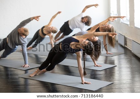 Group of young sporty people practicing yoga lesson with instructor, stretching in Bending Side Plank exercise, Vasisthasana pose, working out, indoor studio image. Wellbeing, wellness concept