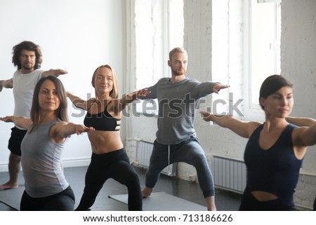 Group of young sporty attractive people practicing yoga lesson with instructor, standing in Warrior Two exercise, Virabhadrasana 2 pose, working out, indoor close up image, studio. Wellness concept