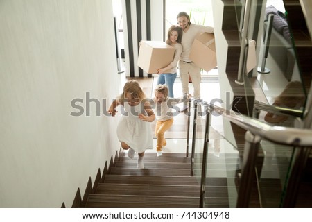 Happy children going upstairs inside two story big house, excited kids having fun stepping walking up stairs running to their rooms while parents holding boxes, family moving in relocating new home