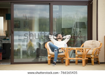 Young calm man relaxing sitting on terrace chair hands behind head, relaxed guy breathing fresh air, meditating with eyes closed outside modern house, resting alone, enjoying pleasant morning outdoor