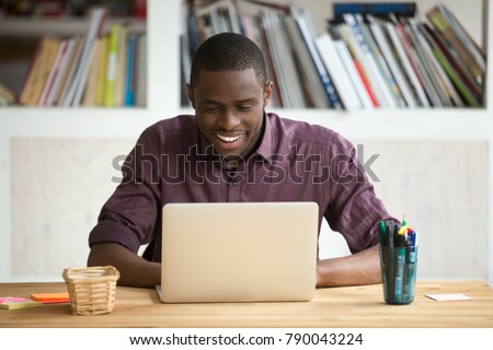 Smiling african-american man using laptop sitting at home office desk, happy excited black e business owner looking at computer screen pleased by reading good news, chatting in social network