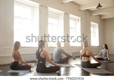 Group of young sporty people practicing yoga lesson with instructor, sitting in Padmasana, Lotus pose, working out, students training in club, loft studio, rear view. Wellness and wellbeing concept