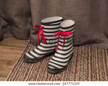 A pair of black and white striped wellington boots with red ribbon bows tied on them. They are standing on a rug with a wooden floor and brown curtain background.