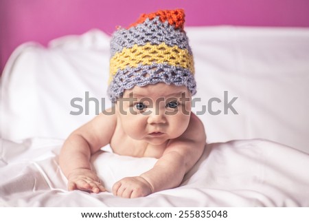 little girl lying on her stomach on the bed, blue eyes, and fun to knit cap on his head, baby cool