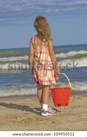 young girl with sand pail at the beach looking out over the water.