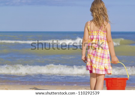 girl standing in front of a large body of water holding a sand pail.