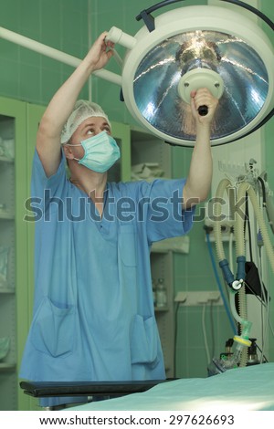 young man surgeon in blue gown, mask and hat directs surgical lamp above the table in operating room