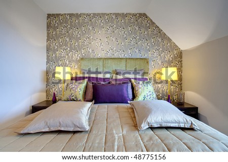 Chic modern bedroom decorated with striking wallpaper dominated by bed dressed with silk duvet, brightly colored cushions and pillows