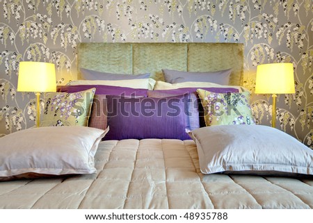 Fashionable bed with silk headboard dressed with brightly colored cushions and pillows, matching duvet, contemporary wallpaper and glass table lamps