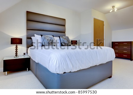 Fashionable modern bedroom simply dressed including bed with oversize headboard, lamp and cushions