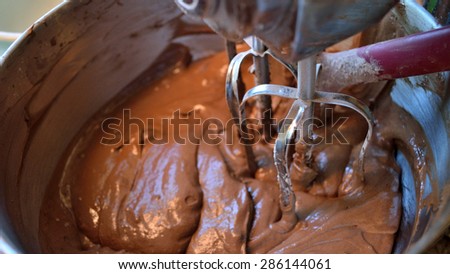 Closeup of chocolate cake mix in stainless still bowl with stainless steel beaters in mix. Cake is mixed and ready to be poured into baking pans. Red spatula in bowl at back.