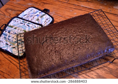 Dark chocolate cake sitting on a cooling rack to cool with colorful oven holders and sea green antique bowl with cake mix in it background on distressed wooden table in old-fashioned kitchen.