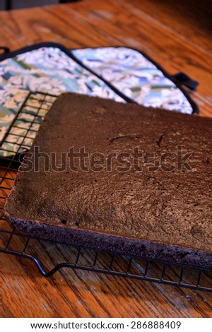 Dark chocolate cake sitting on a cooling rack to cool with colorful oven holders in background on distressed wooden table in old-fashioned kitchen.