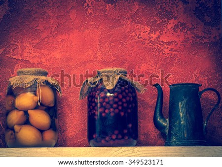 Winter stocks of vitamins in pantry -- home canned food in glass jars, compotes of cherries and lemons. Stewed fruit and old jug in a cellar - still life in retro style on red background.