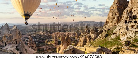 Wide panorama of Cappadocia landscape - colored balloons flying over mountain peaks and fantastic valley of ancient cave town Uchisar. Air balloons above Turkish National Park in Goreme.