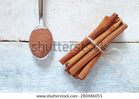 Some cinnamon sticks tied with a natural rope. Next, a spoon with some powdered cinnamon. All on a white wooden table. Top view