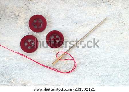 Sewing needle threaded with a red thread ready for sewing. Next , three buttons. Scene on a white wooden table . Top view.