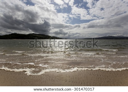 Ireland west coast landscape scene during stormy weather in summer on a sandy beach with small waves breaking and sun shining through broken cloud.