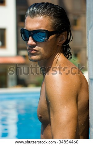 Young sexy muscular man standing in swimming pool in sunglasses
