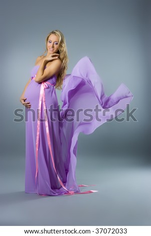 Beautiful young pregnant blonde womanin lilac dress in studio shot on gray background