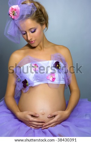 Young beautiful pregnant woman in violet dress and bonnet with flower in studio shot on gray background
