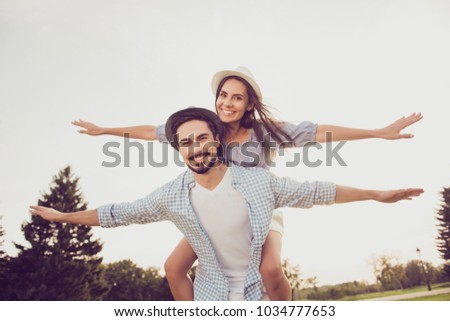Cute romantic bearded brunet in checkered shirt, dreamy lady rides him on rear. Leisure, chill happiness, lawn stroll, relax, romance lifestyle, well dressed in blue, partners posing in headwear
