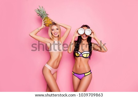 Portrait of hot, charming, stylish, pretty, trendy, crazy ladies, tourists in swim suits, blonde having ananas on her head, brunette having coconut on eyes\' place, standing over pink background