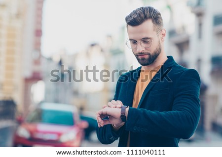 Portrait of smart responsible man with modern hairdo looking at watch on wrist over blurred street background hurry up for meeting. Management employment job concept
