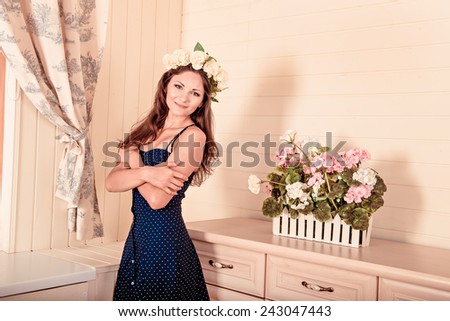 girl stands near a white dresser with a wreath on head. there are flowers on the dresser.