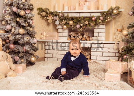 at Christmas little boy smiling.baby sits near a Christmas tree with gifts. little gentleman sitting near the white fireplace