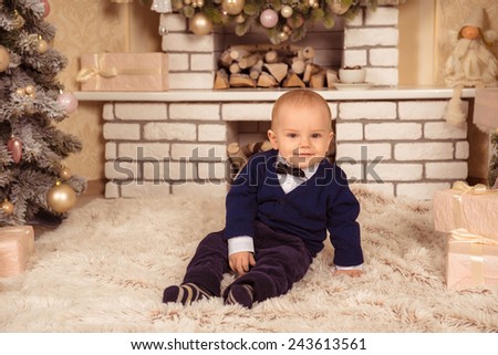 at Christmas little boy smiling.baby sits near a Christmas tree with gifts. little gentleman sitting near the white fireplace
