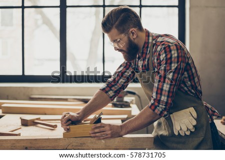 side view photo of stylish handsome cabinetmaker work with jointer and plank. Man with brutal hairstyle, beard and glasses.