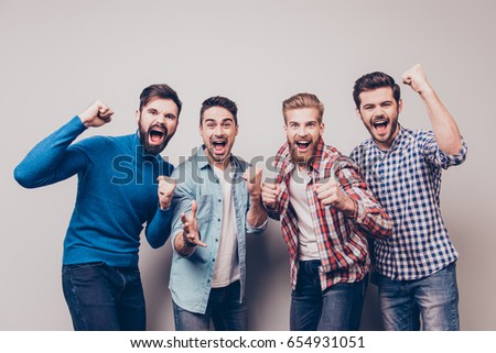 Yes! Winners! Four cheerful young men are standing and gesturing for the victory on pure background in casual outfit and jeans. They are fans of sports games as football, basketball, hockey, baseball