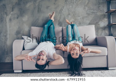 Go crazy together! Relaxing upside down is fun. Cheerful brother and sister are lying on the beige sofa at home indoors, in jeans and white casual t shirts, gesturing