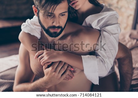 Cheating, jealous, obsession, possession concept. Handsome sad half naked young guy is looking down, thinking about cheating to his wife with gorgeous lover, who hugs him from the back, cropped photo