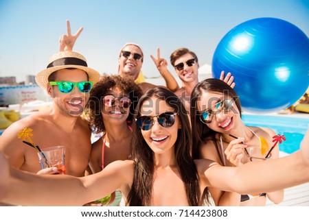Selfie time! Six cheerful friends in fashionable different swim wear and sun protecting eyewear, caps, with drinks are posing for a selfie photo, that brunette lady is taking, beaming smiles, sunshine