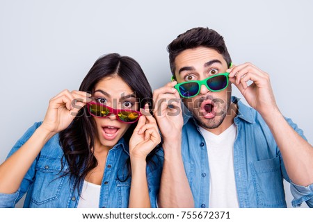 Concept of large sales and discount. Close up photo of two excited and wondered people with open mouths dressed in casual clothes, they are touching colorful glasses, isolated on grey background