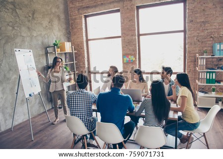 Young smart lady in glasses is reporting to the team of colleagues about the new project at the meeting with the white board. Workers are listening to her, all dressed in casual outfits