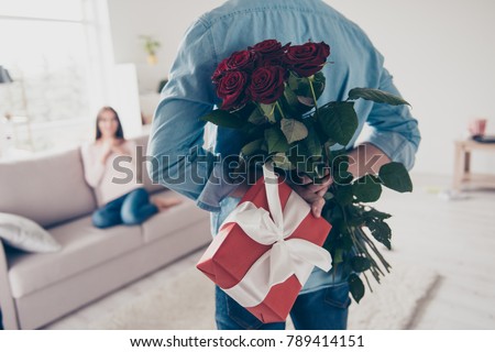 Unexpected moment in routine everyday life! Cropped photo of man\'s hands hiding holding chic bouquet of red roses and gift with white ribbon behind back, happy woman is on blurred background