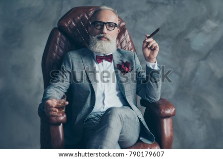 Cool man in glasses, hold cigarette,  glass with brandy, in formal wear, tux with red bowtie and pocket square, sit in leather chair over gray background, looking to the camera, shares, stock, money