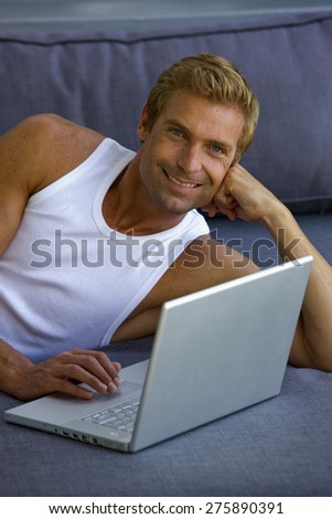Beautiful young blond man lying down on blue sofa and using laptop.